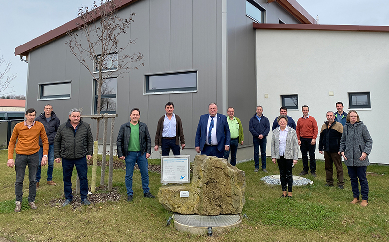 Gruppe von Personen steht im Freien vor einem modernen Gebäude; im Vordergrund ein Gedenkstein mit Tafel auf einer Rasenfläche, umgeben von Bäumen und Gehwegen. Geben Sie Ihr Feedback zu BizChat ab.