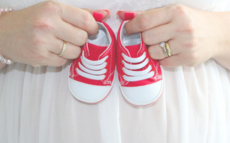 Zwei Hände halten ein Paar kleine rote Babyschuhe vor einem hellen Stoffhintergrund, als symbolische Darstellung von Nachwuchs.