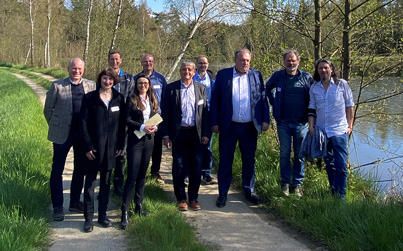 Eine Gruppe von Erwachsenen geht gemeinsam einen schmalen Weg entlang eines Flusses durch eine grüne, bewaldete Landschaft.