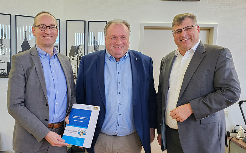 Drei Männer in einem Büro; der Herr links hält eine Dokumentenmappe des Beauftragten für Bürokratieabbau in den Händen