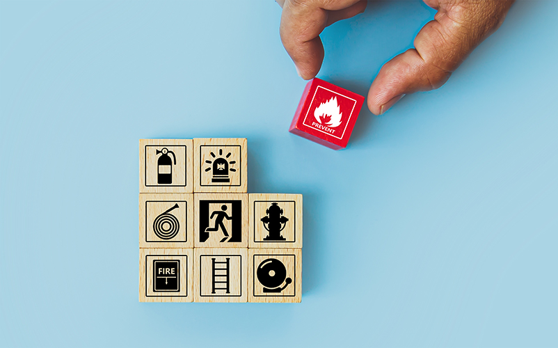 Eine Hand platziert einen roten Würfel mit Flammen‑Symbol über mehreren Holzklötzen, die Sicherheits‑ und Brandschutzpiktogramme zeigen.