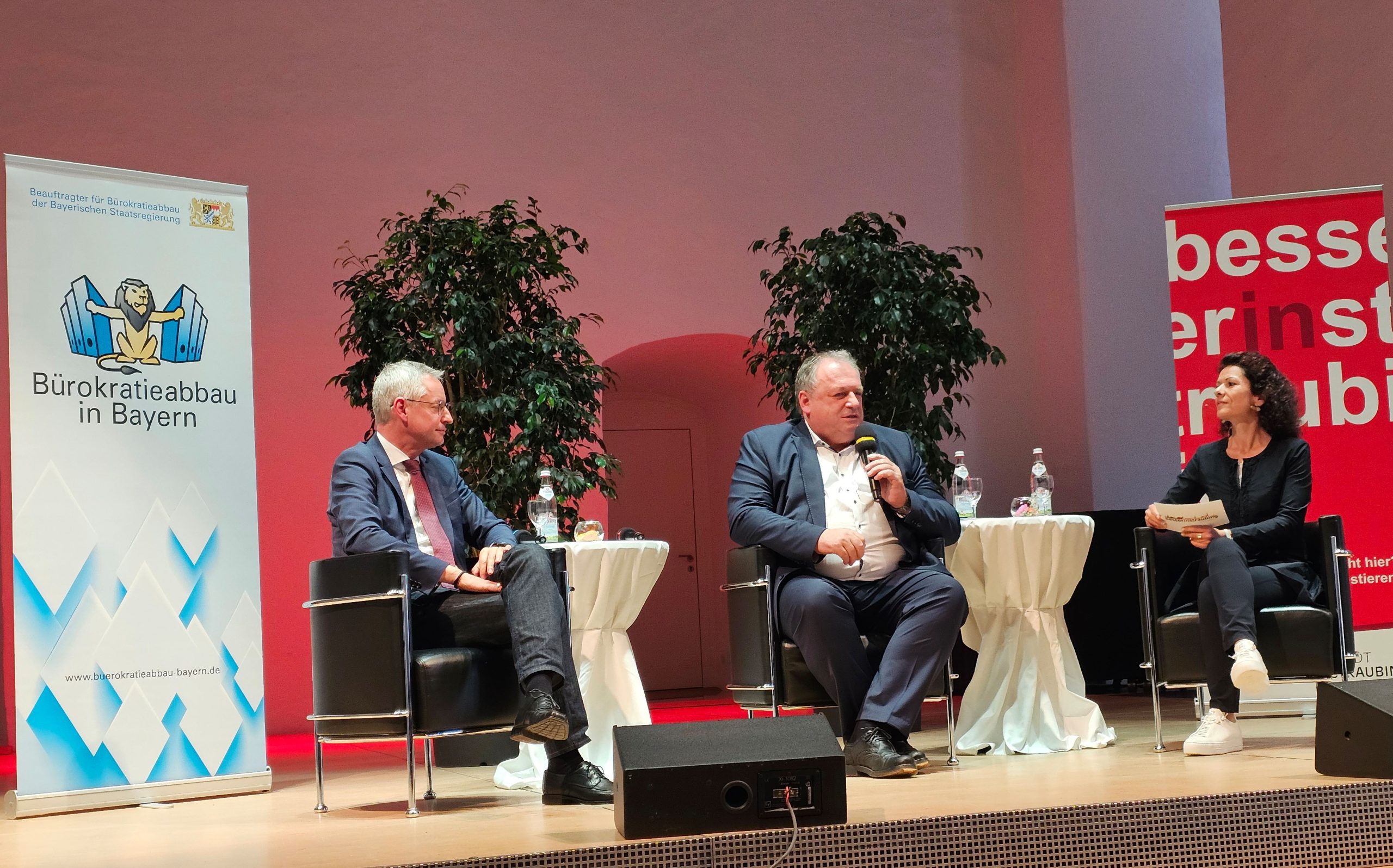 Zwei Männer und eine Frau auf einem Podium sitzend flankiert von zwei Rollups