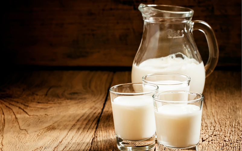 Eine Glaskaraffe und drei Gläser gefüllt mit Milch stehen auf einem Holztisch.