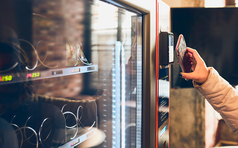 Eine Person hält ein Smartphone an das Kartenlesegerät eines Snack- und Getränkeautomaten, um kontaktlos zu bezahlen.