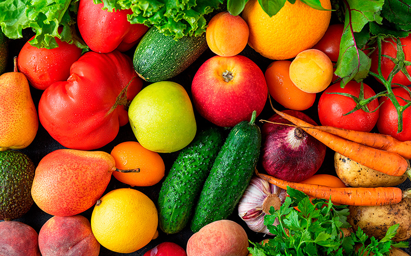 Verschiedene frische Obst- und Gemüsesorten wie Äpfel, Gurken, Tomaten, Karotten, Paprika und Salat liegen bunt gemischt nebeneinander.