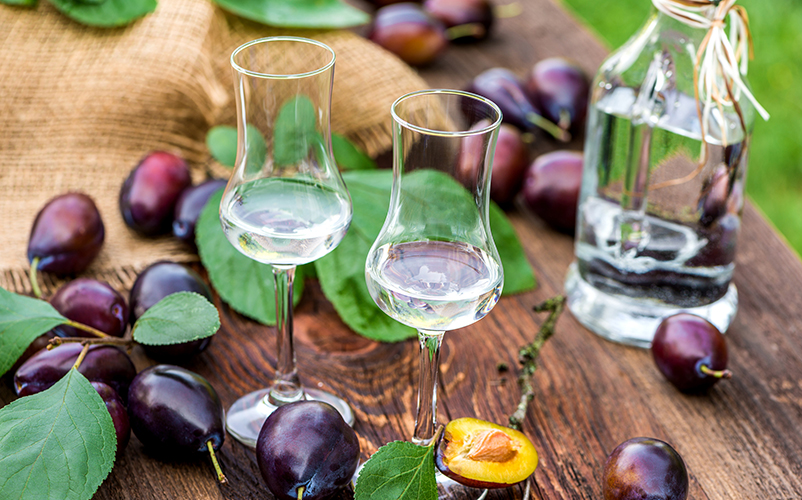 Zwei Gläser mit klarem Pflaumenschnaps stehen auf einem Holztisch, umgeben von frischen Pflaumen und einer Flasche Schnaps.