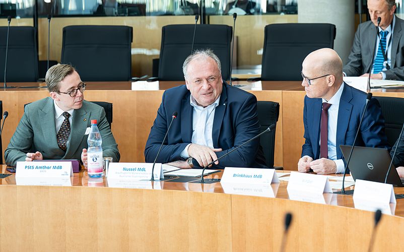 Mehrere Personen sitzen an einem Konferenztisch mit Mikrofonen und Namensschildern und führen ein Gespräch in einem Sitzungssaal.