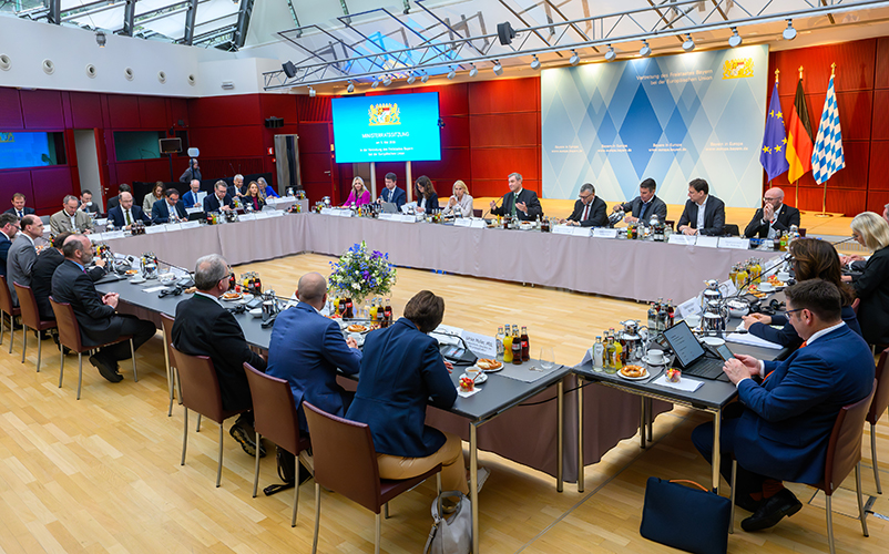 Kabinettssitzung in einem Konferenzsaal mit U-förmiger Tischordnung, zahlreichen Teilnehmenden, Leinwand sowie Bayern- und EU-Flaggen im Hintergrund.